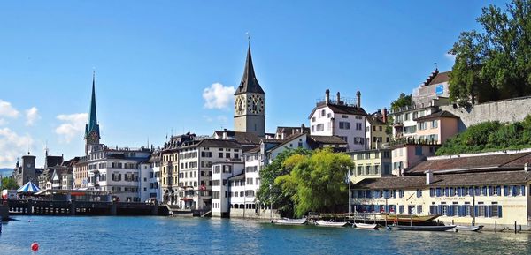 Churches and buildings by limmat river in city against blue sky