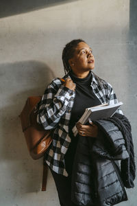 Thoughtful woman holding book and jacket standing with backpack against gray wall