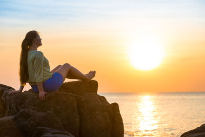 Man sitting on rock by sea against sky during sunset