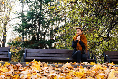 A short-haired woman wraps herself in a scarf sitting on a bench in an autumn park against trees