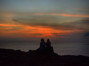 Rear view of couple sitting on coast against cloudy sky at dusk