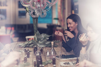 Smiling woman smelling perfume bottle while sitting by colleague at table in workshop seen from window glass