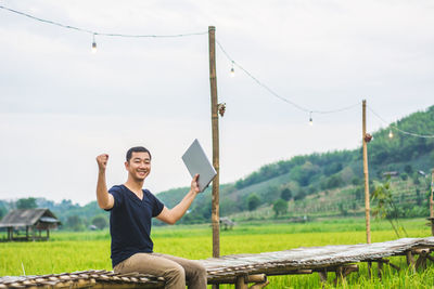 Man with arms outstretched holding laptop while sitting on boardwalk at rice paddy