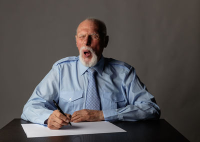 Senior businessman writing in paper and making face while sitting on table against gray background