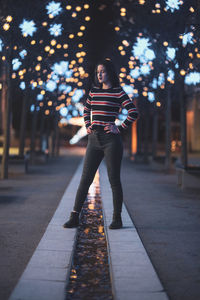 Woman standing on footpath against illuminated lights at night
