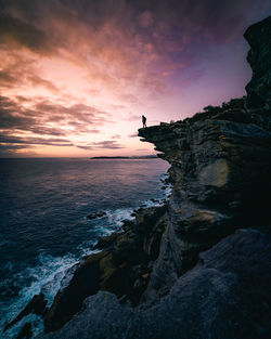 Man on cliff by sea against sky during sunset