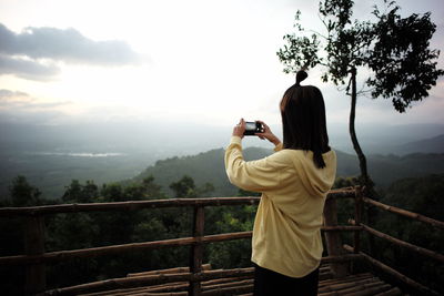 Side view of woman photographing while standing on railing against sky during sunset