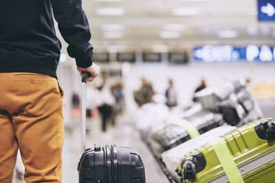 Midsection of young man with luggage standing by conveyor belt at airport