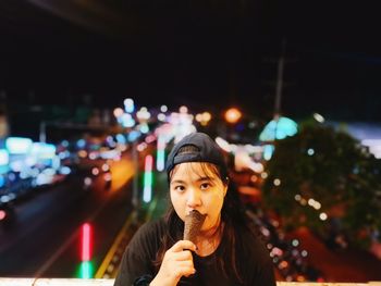 Woman eating ice cream standing against illuminated city street at night