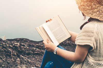 Midsection of woman reading book while sitting on rock by lake