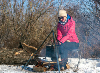 Woman with sunglasses by campfire, pot of soot over bonfire on tripod, winter cooking at camping