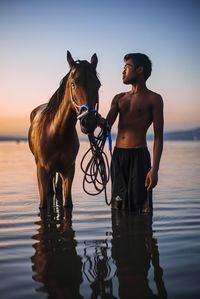 Man with horse in sea against sky during sunset