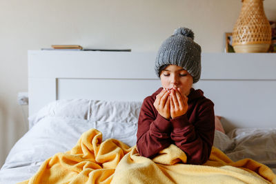 Child wearing grey warm hat sits on the bed under warm blanket and tries to warm up her hands