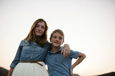 Low angle portrait of siblings standing against clear sky during sunset