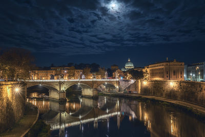 Illuminated bridge over river by buildings against sky at night