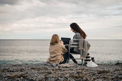 Women sitting on shore at beach against sky