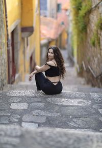 Full length of young woman sitting on sidewalk in city