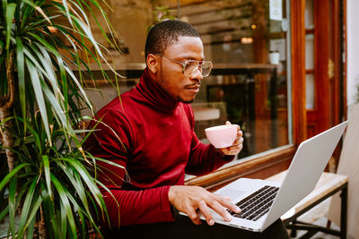 Side view of rich stylish focused african american male entrepreneur sitting on bench near cafe outdoors and browsing laptop while drinking delicious coffee and working remotely on project