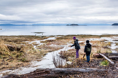 Full length of friends standing at lakeshore against cloudy sky