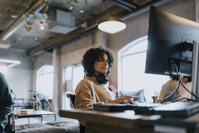 Female hacker with wireless headphones around neck using computer while working in office