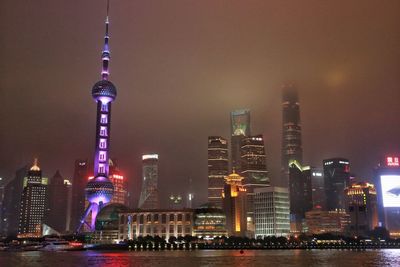 Low angle view of illuminated oriental pearl tower in front of river against sky in city