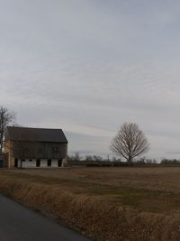 Bare trees on field by houses against sky