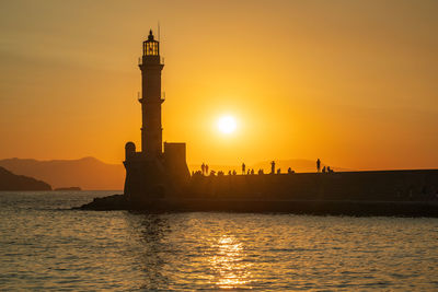 Lighthouse by sea, people gathering together against sky during sunset