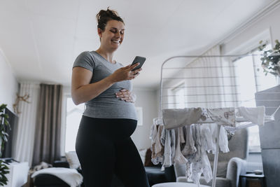 Pregnant woman using phone while doing housework and hanging baby clothes on drying rack