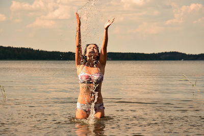 Woman splashing water while standing in lake against sky during sunset