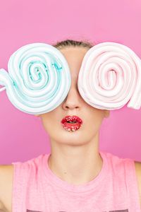 Close-up of woman holding lollipops as eyes while standing against pink background