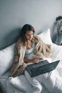 Sick young woman using laptop on bed against wall at home