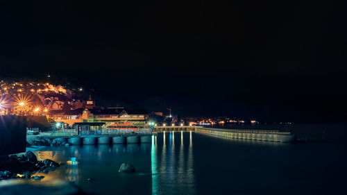 Illuminated city by sea against sky at night