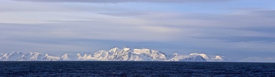 Panoramic view of snowcapped mountains in front of sea against cloudy sky