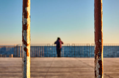 Rear view of woman standing on rusted metal post and boardwalk at sea shore against clear sky