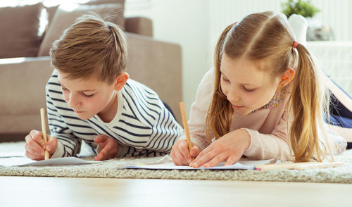 Siblings writing on paper while lying at home