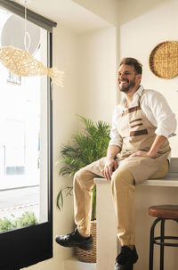 Delighted male cook in apron sitting on counter in modern asian restaurant and looking away