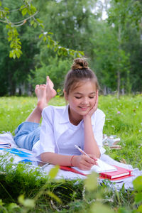 A young schoolgirl lies on the grass in the park with a notebook and a pen.