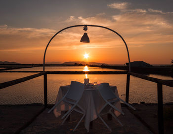 Empty table and chairs by sea against sky during sunset