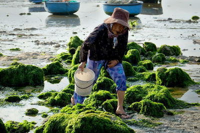 Senior woman holding bucket while working on mossy rocks at beach