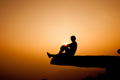 Silhouette of woman sitting on cliff against sky during sunset