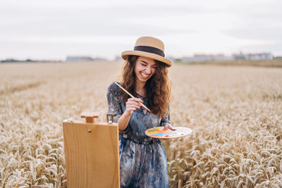 A young woman with curly hair and wearing a hat is painting in nature. 