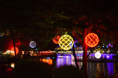 Illuminated ferris wheel by river against sky at night