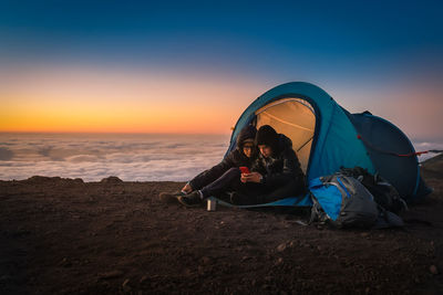 Brothers using mobile phone while camping on mountain against sky during sunset