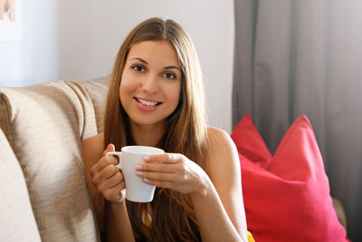 Portrait of young woman holding coffee cup while sitting on sofa at home