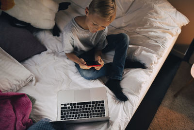 High angle view of boy using mobile phone sitting with laptop on bed at home