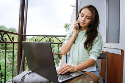 Young woman sits on a balcony at a table working with a phone behind a laptop