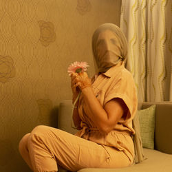 Woman face covered with fabric holding flower while sitting on sofa