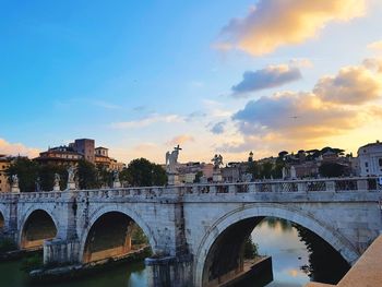 Bridge over river by buildings against sky during sunset