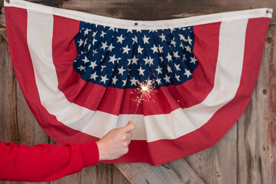 Cropped hand holding lit sparkler against american flag on wall