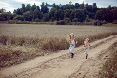 Two girls in long dresses running along the path to the field of dry wheat